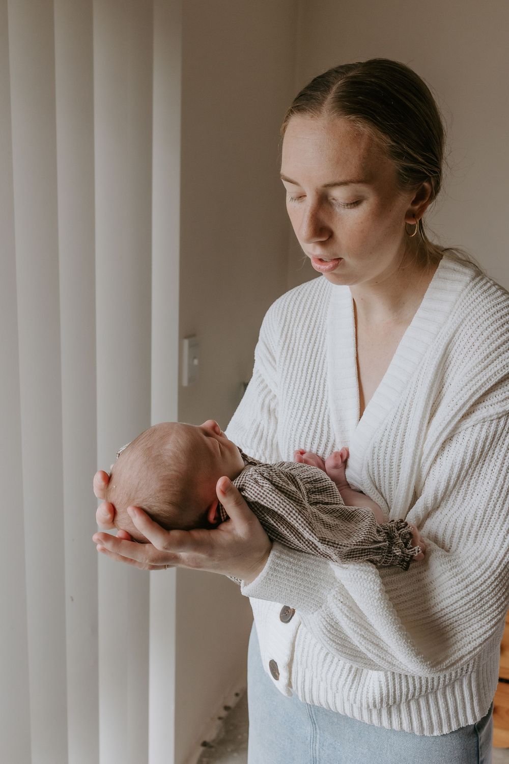 A young mother in a white cardigan tenderly holds her sleeping newborn baby near a bright window.