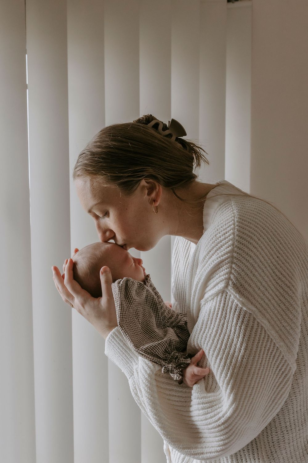 A mother in a white knit sweater gently kisses her newborn baby near bright white window blinds.