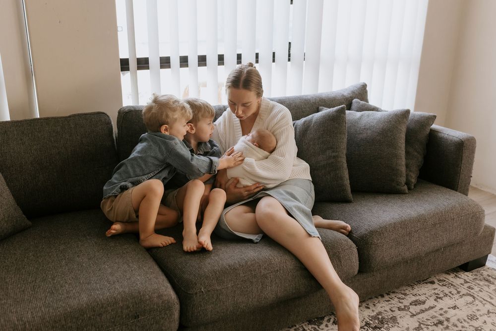 A mother sits on a gray sofa with her two young children, cradling a newborn baby in a bright living room.