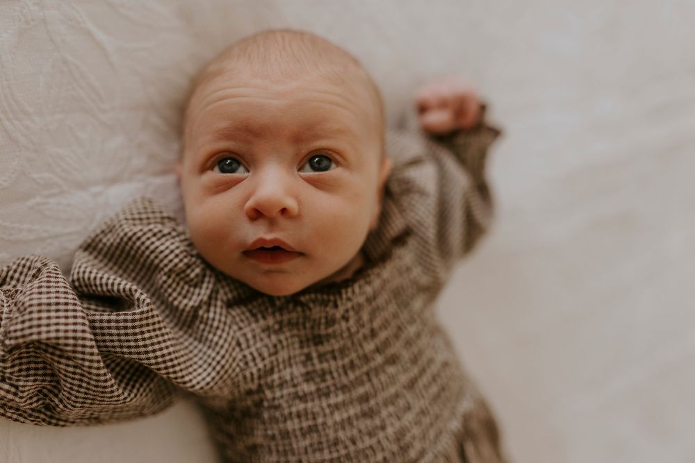 Newborn baby in a brown knit romper lying on a white blanket, looking up with wide eyes and one arm raised.