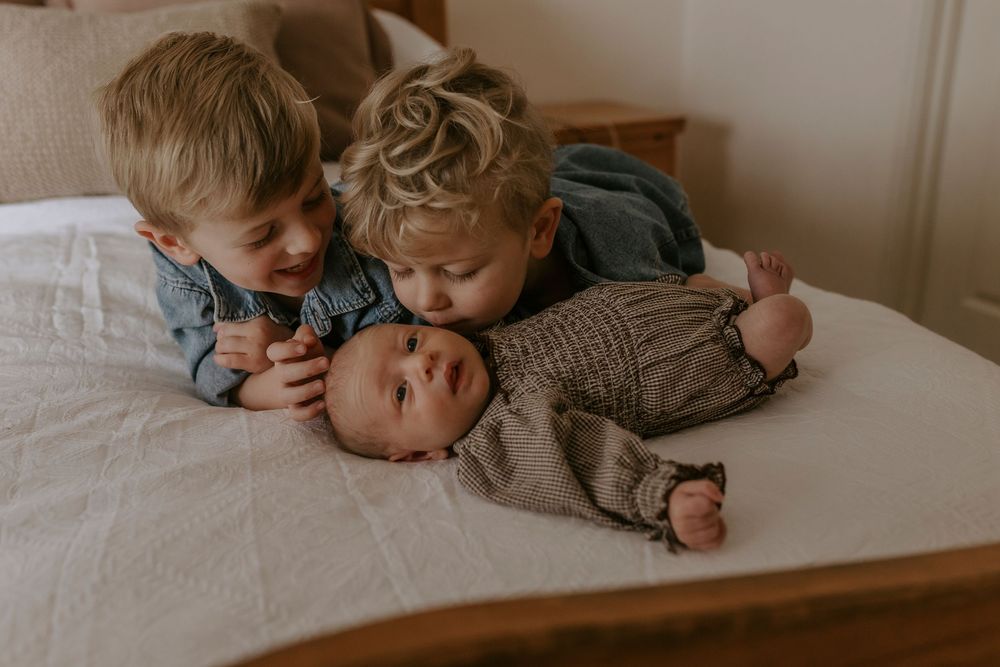Two young boys cuddle with their newborn sibling on a white bed in a sweet family portrait.
