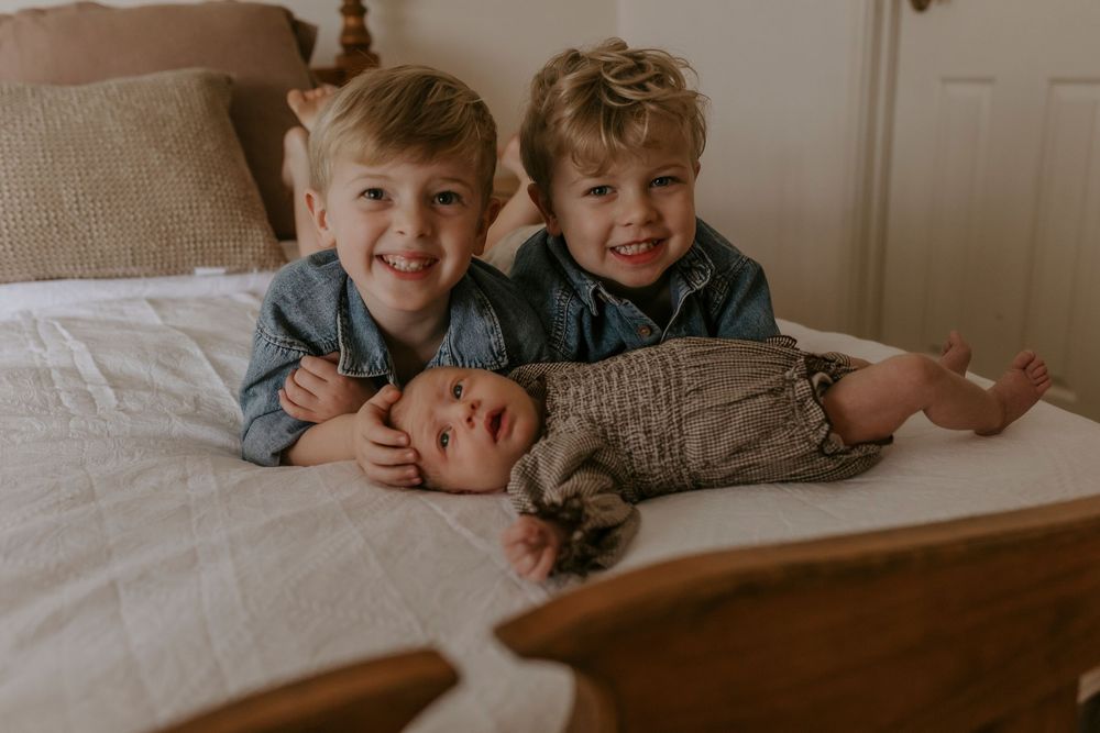 Two smiling boys lying on a bed with their newborn sibling in a cozy bedroom family photo.