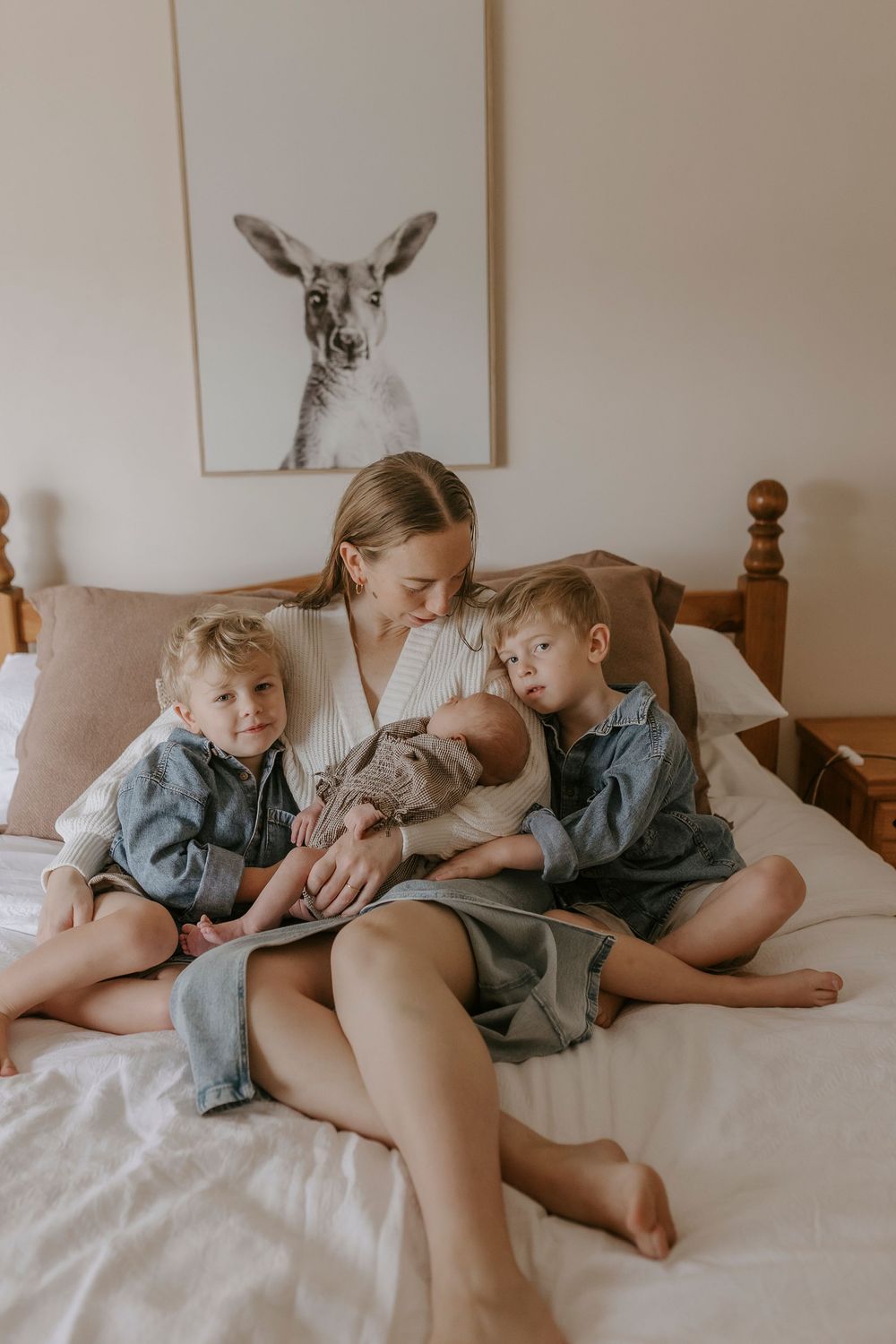 Mother sits on bed holding newborn baby flanked by two toddlers in denim, with kangaroo wall art behind.
