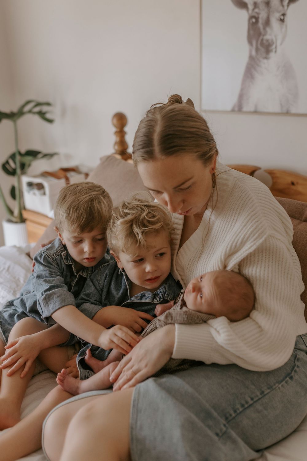 Mother sitting on bed holding newborn baby while two young toddler siblings lean in close to look.