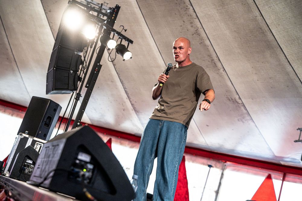 Bald man in olive t-shirt speaking into microphone on stage at outdoor festival event.