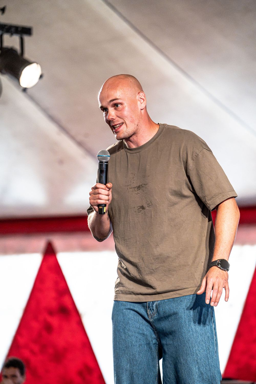 Bald man in olive t-shirt and jeans speaking into microphone on outdoor stage with red tent backdrop.