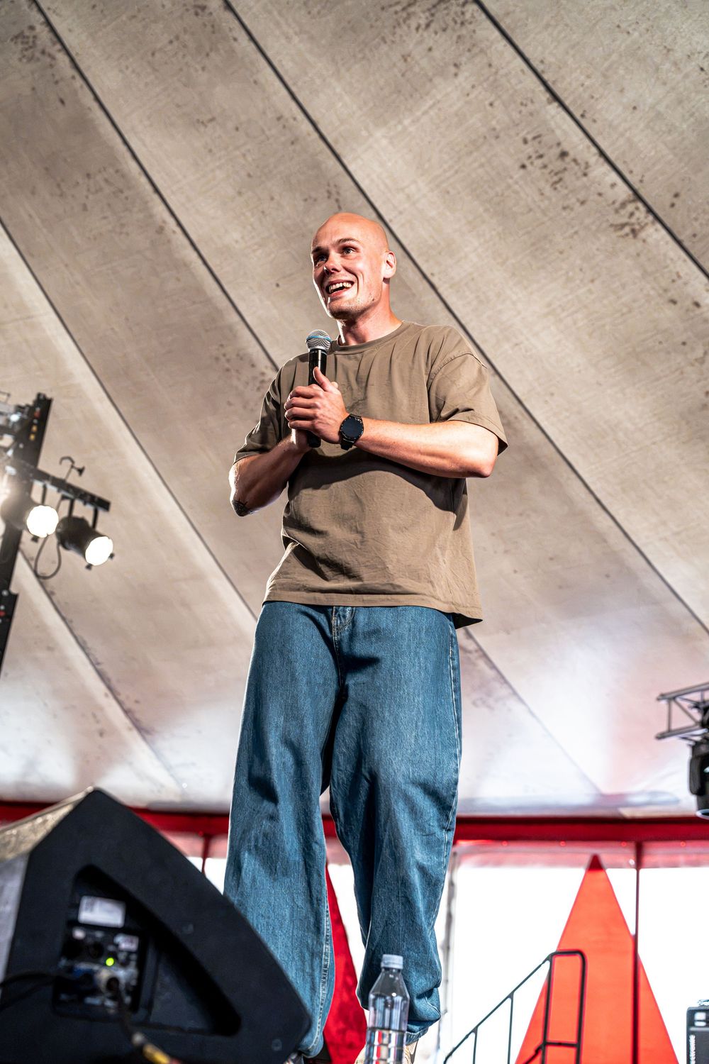 Bald male speaker holding microphone on stage wearing olive t-shirt and jeans performing at outdoor event.