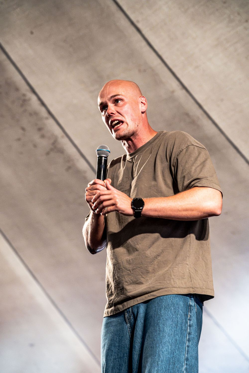 Bald man speaking into microphone on stage wearing olive green t-shirt and jeans at live event.