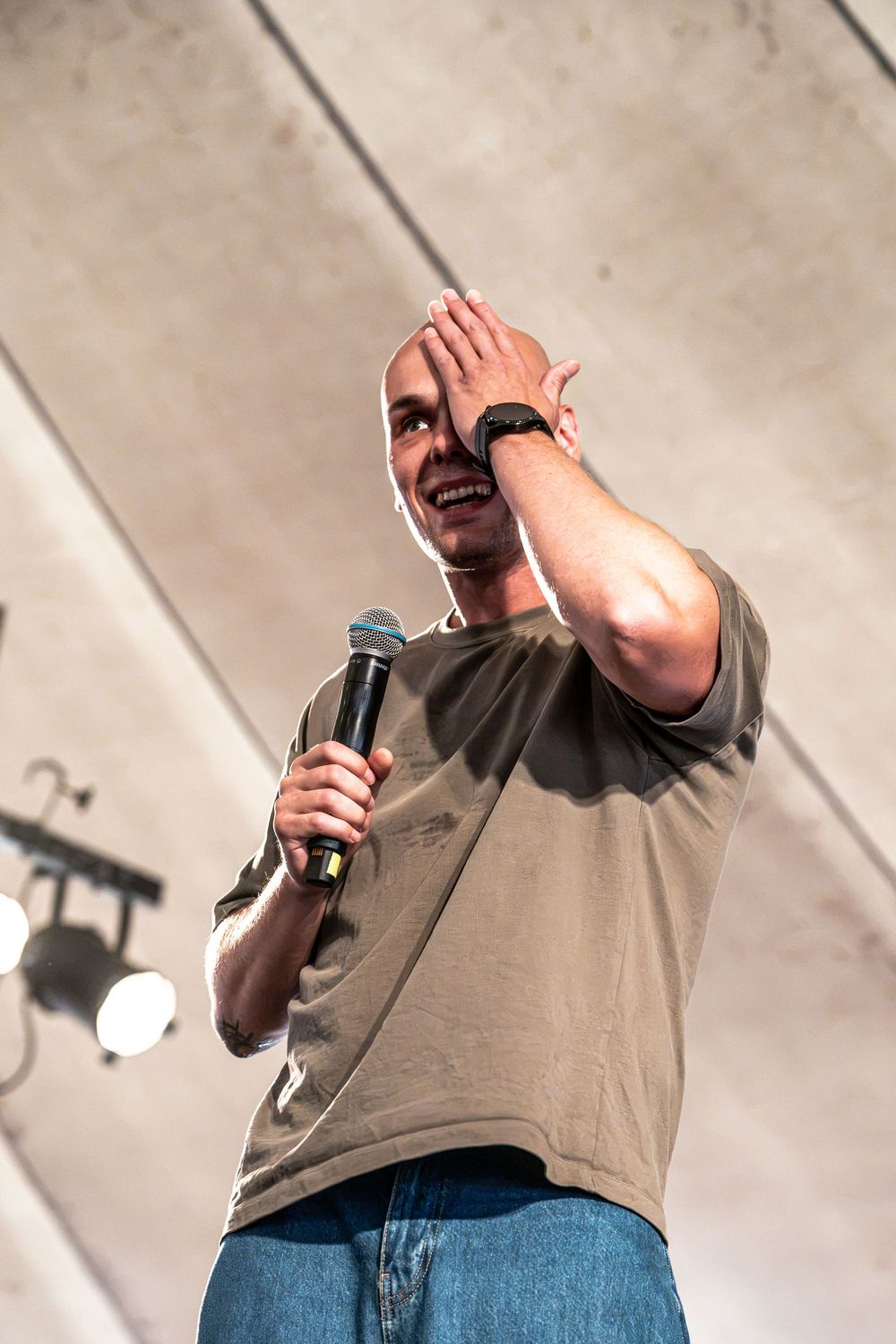 Bald man smiling on stage holding a microphone, raising his hand to his forehead, wearing a gray t-shirt and jeans.