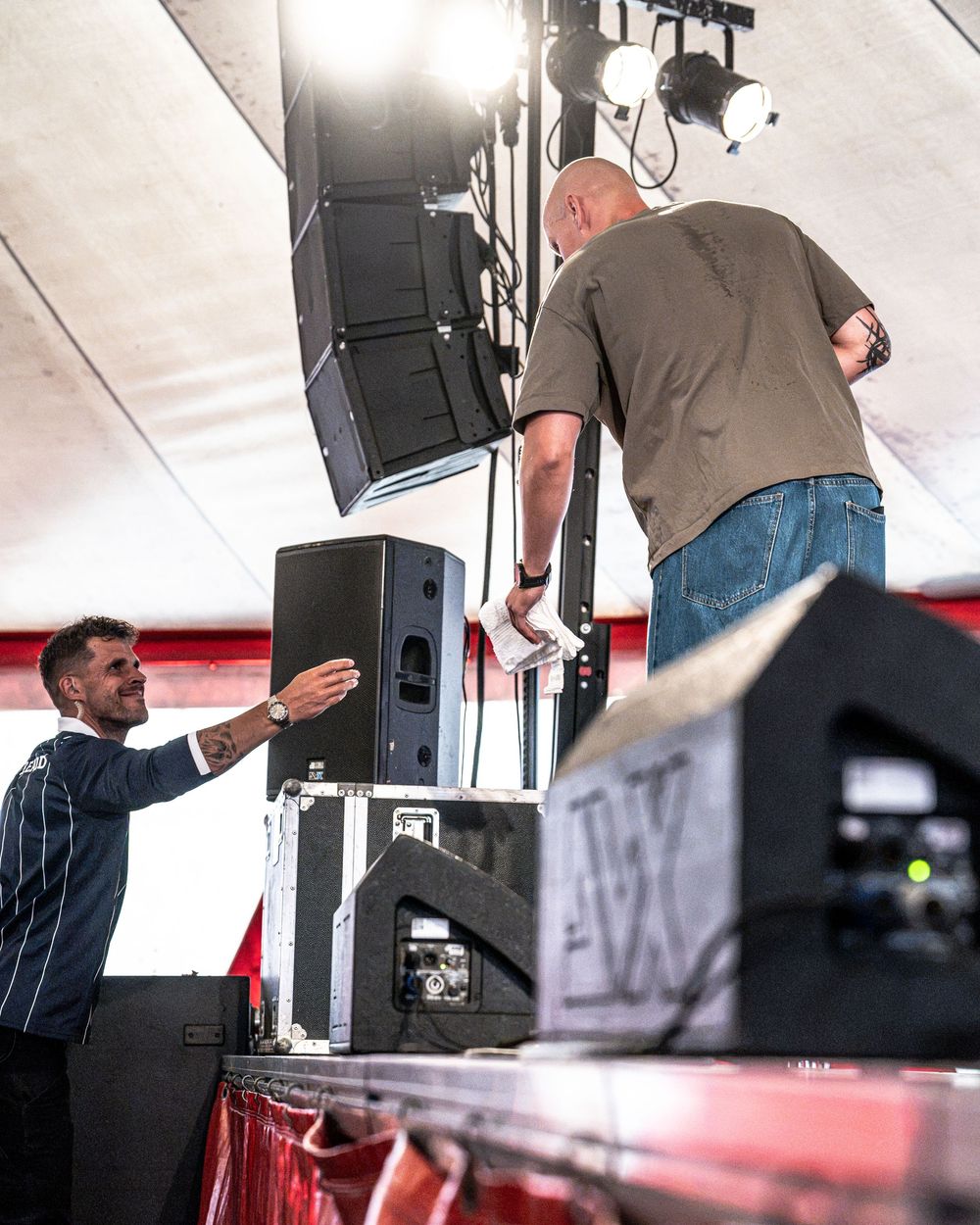 Two men set up large black speakers on a stage under a white tent at an outdoor event.