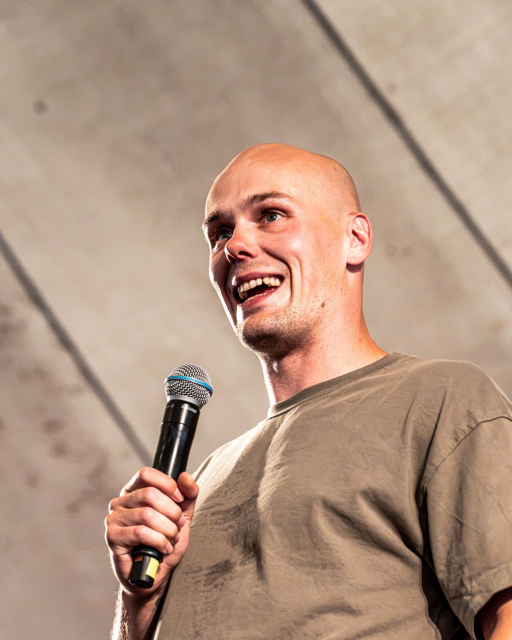 Bald man smiling and holding a microphone while speaking on stage in a tan t-shirt.