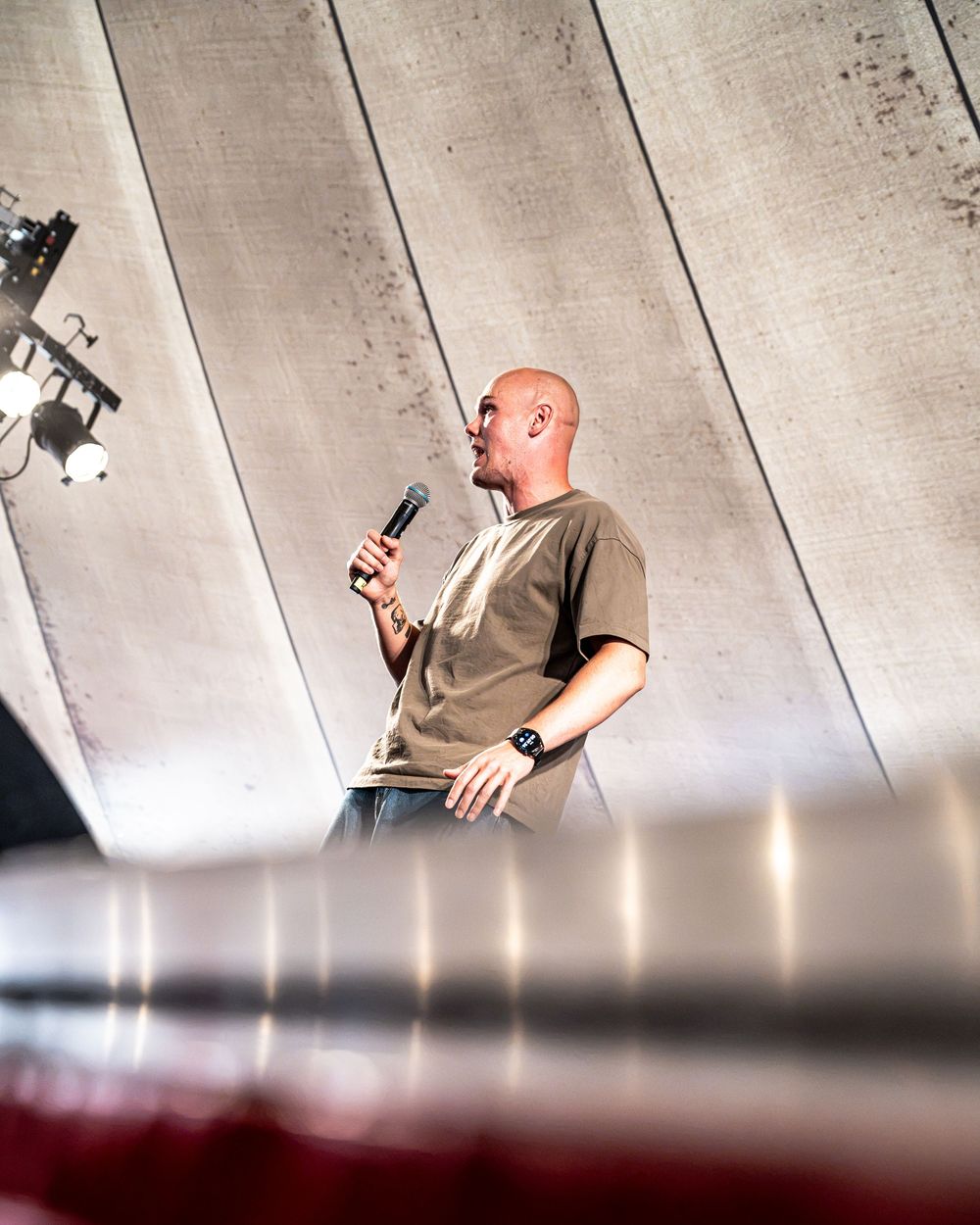 Bald male speaker holding microphone on stage with dramatic lighting and metallic backdrop.