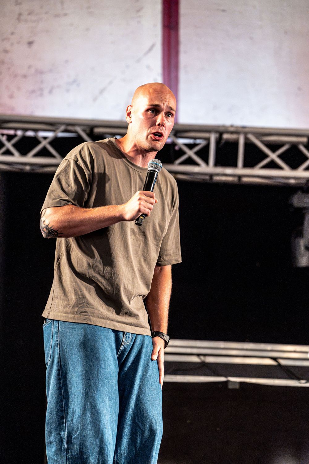 Bald male speaker performing on stage holding microphone under stage lighting.