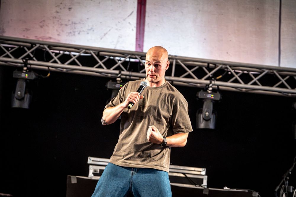 Bald man in olive shirt performing on stage with microphone at indoor venue with truss lighting.