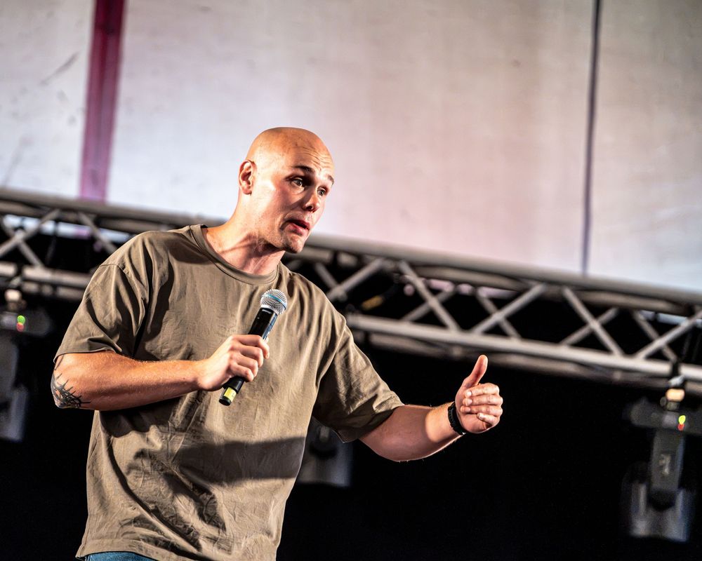 Bald man in olive t-shirt speaking on stage holding a microphone with a thumbs up gesture.