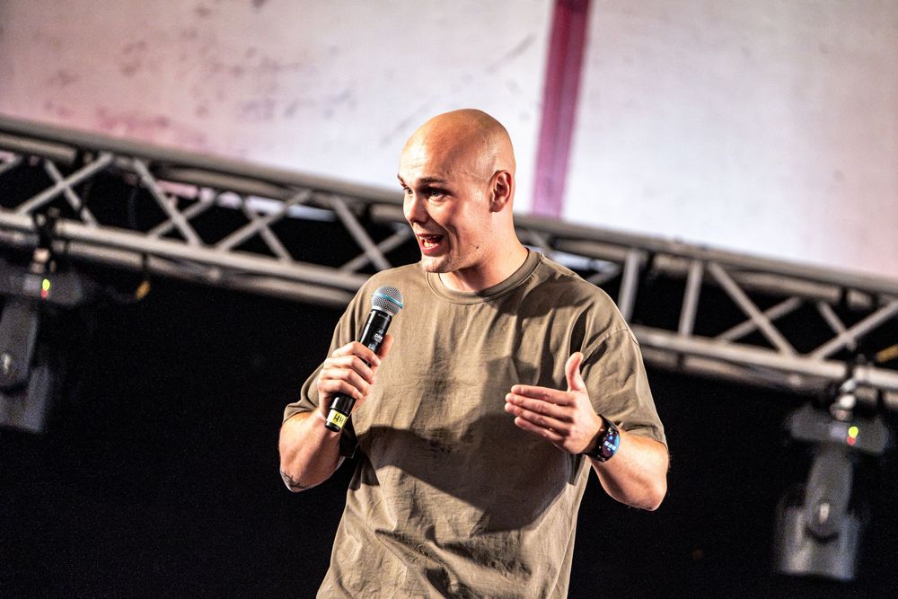Bald male performer smiling on stage holding microphone in olive t-shirt at live music event.