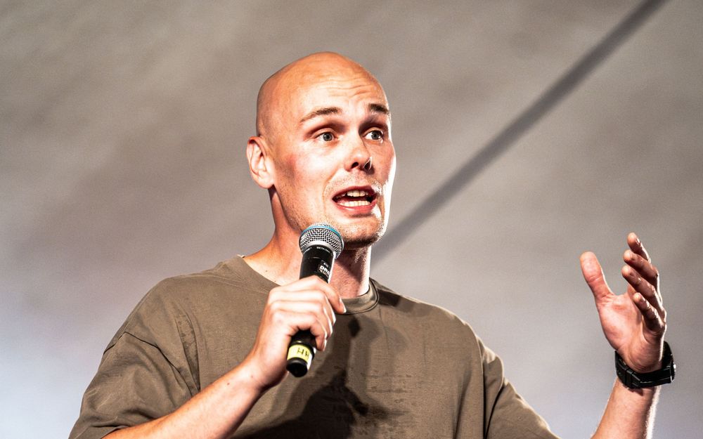 Bald man speaking into microphone on stage, gesturing with hand during live presentation or public speech.