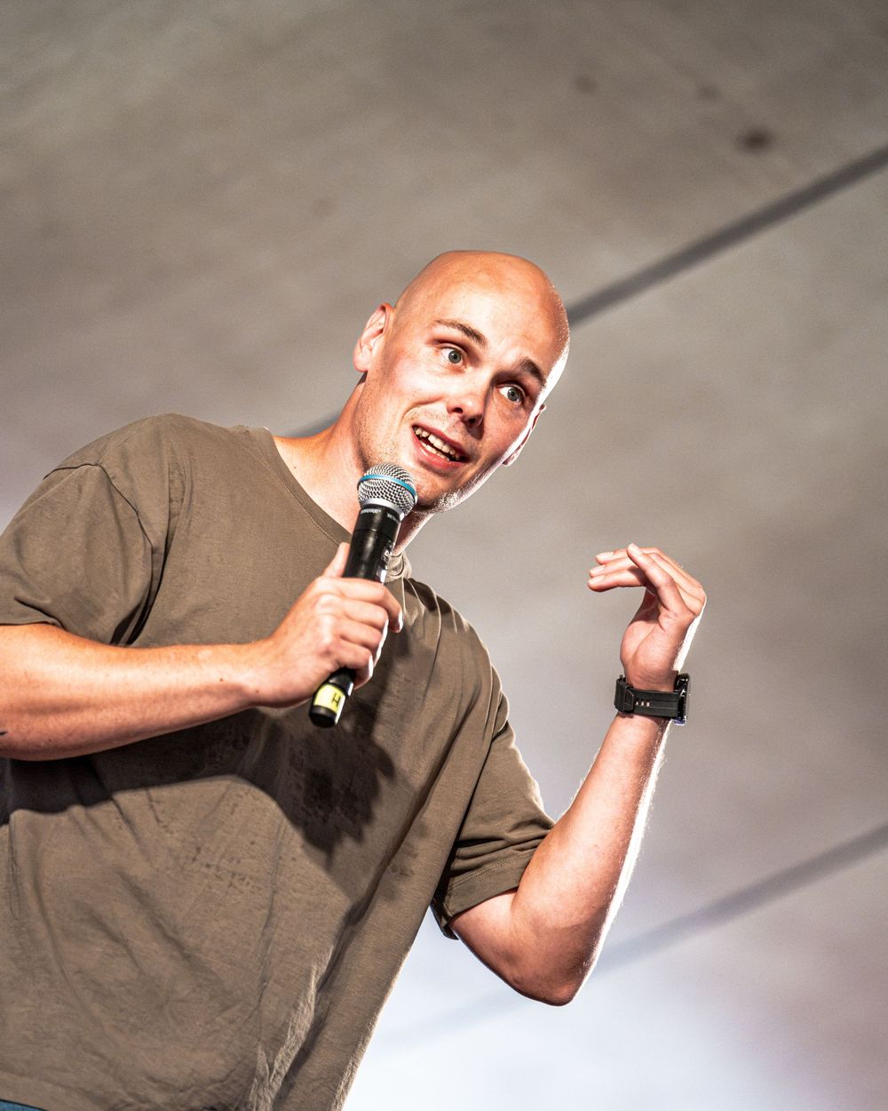Bald male performer gesturing while holding microphone on stage in olive t-shirt at outdoor venue.