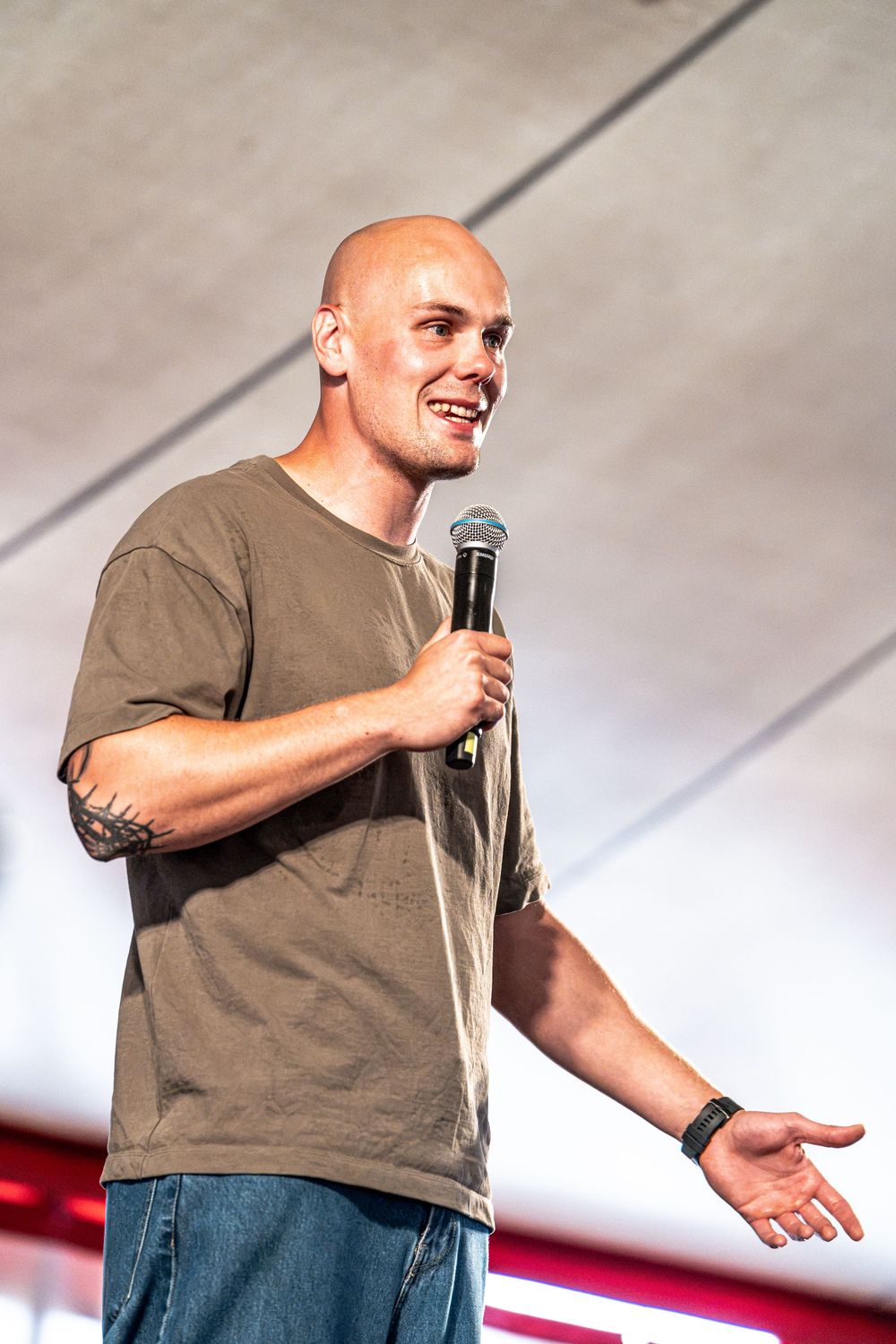 Bald man with tattoos speaking into a microphone on an outdoor stage wearing a khaki t-shirt.
