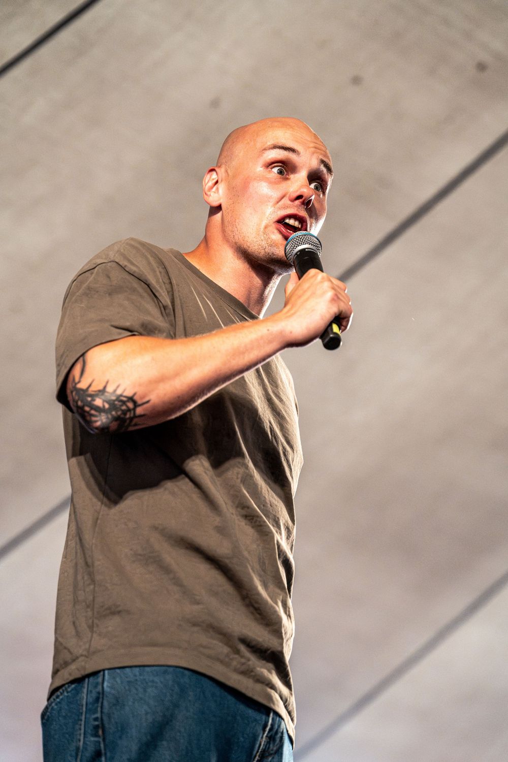 Bald male performer with tattoos singing into microphone on stage wearing olive t-shirt and jeans.