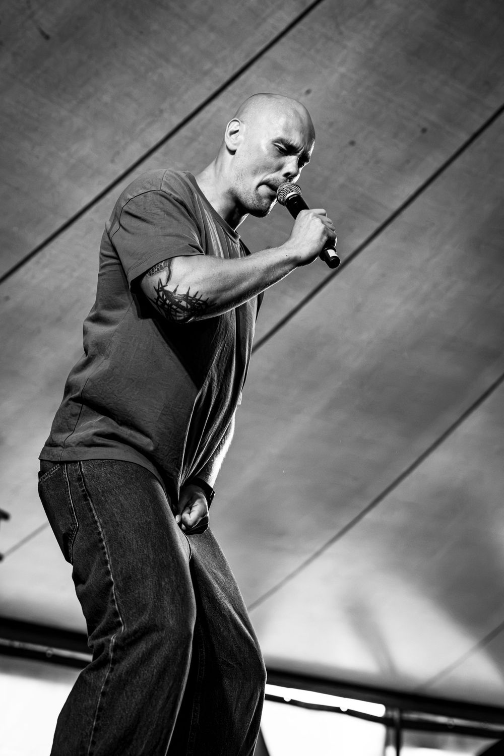 Bald male singer performing on stage, leaning forward into microphone, wearing jeans and t-shirt, black and white photo.