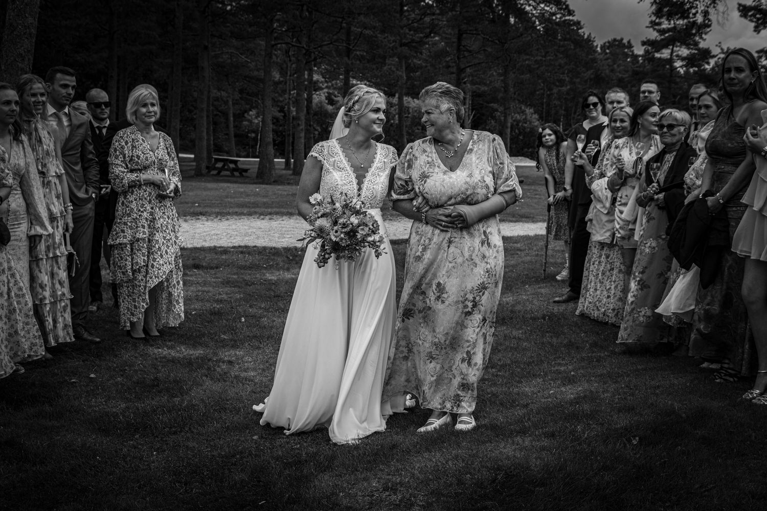 Black and white photo of two women in elegant dresses laughing together at an outdoor wedding ceremony.