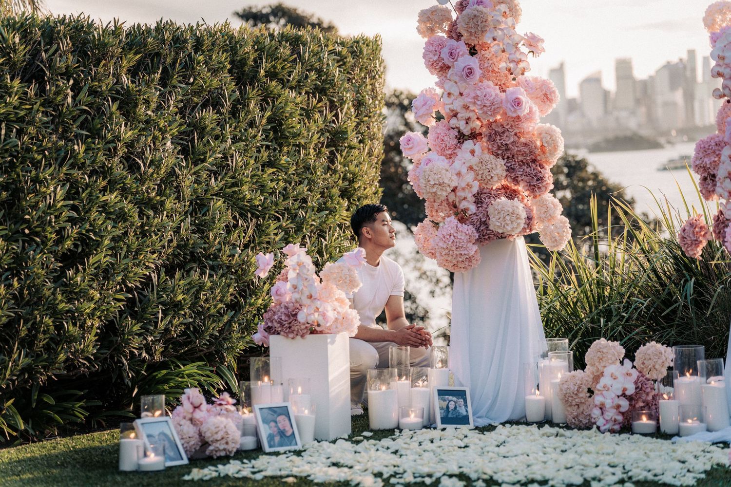 Romantic outdoor proposal setup with pink floral arrangements, candles, framed photos, and city skyline backdrop.