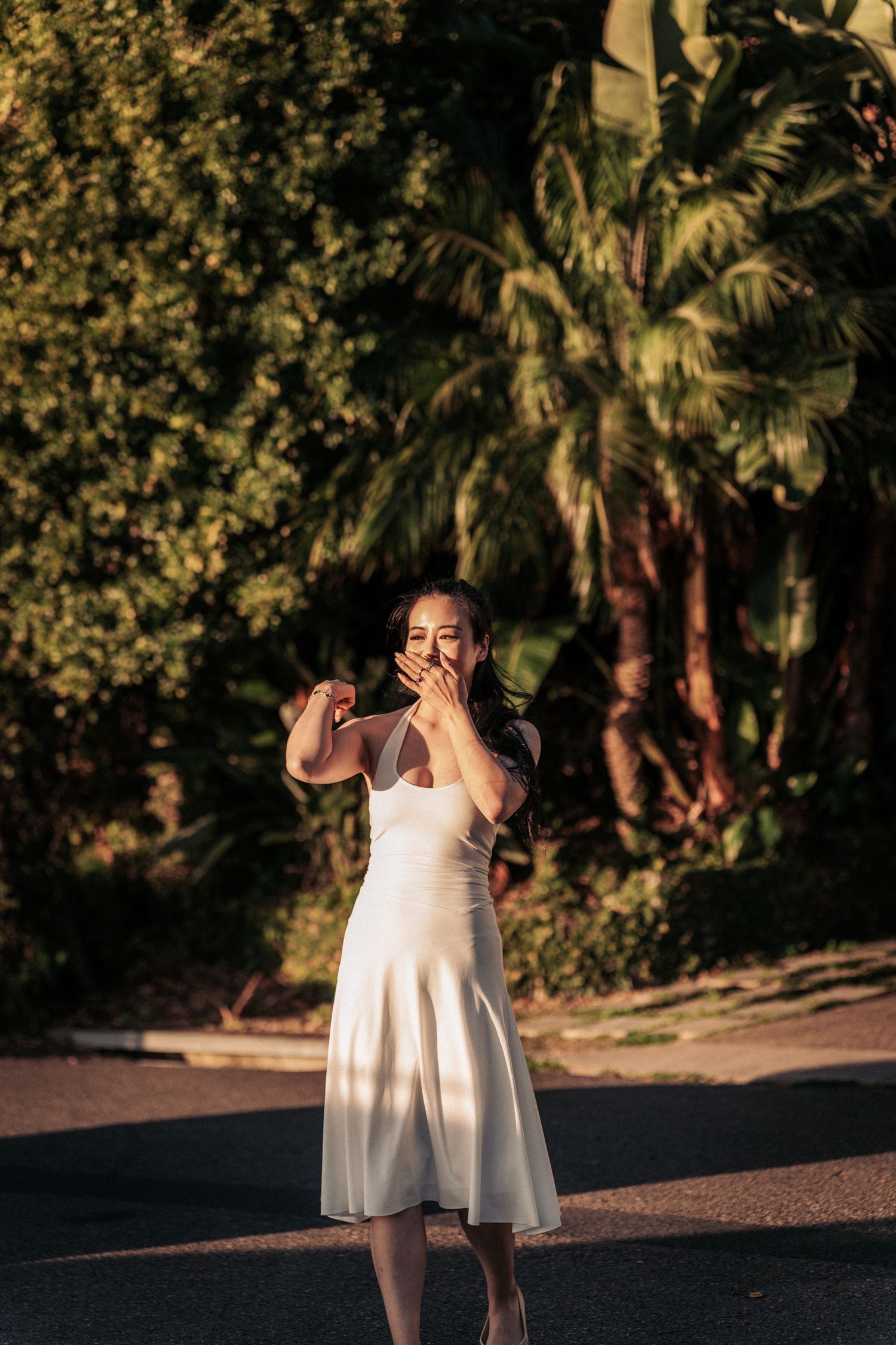 Woman in white midi dress covering mouth with hand, standing on sunlit street with lush tropical palm trees behind her.