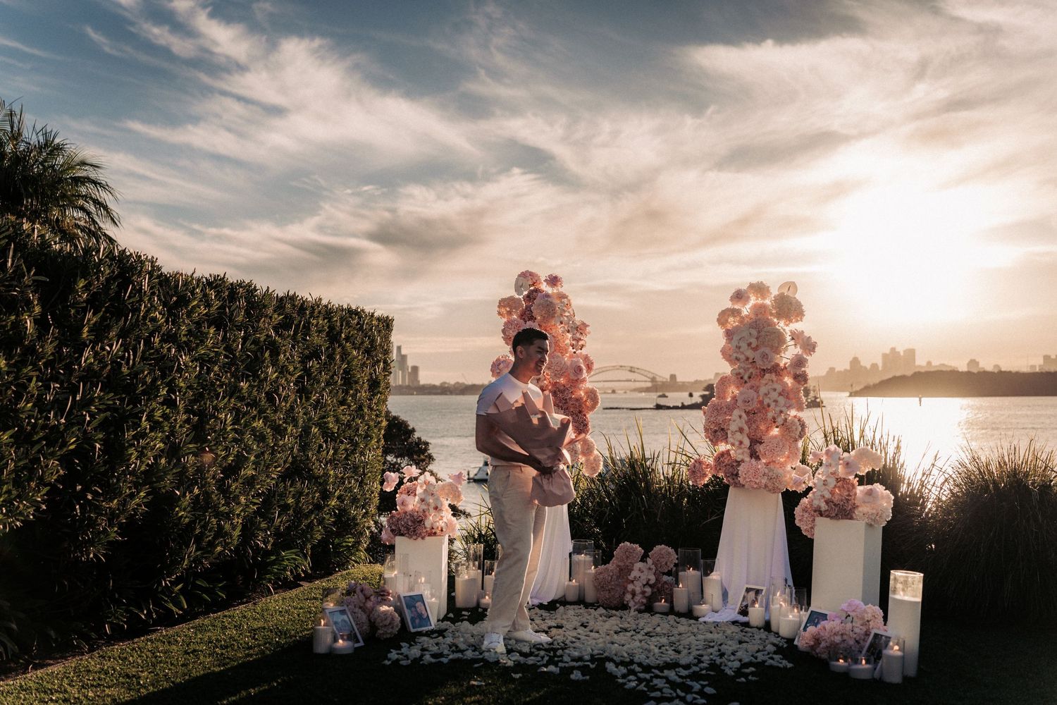 Couple embraces at sunset proposal setup with pink floral arrangements and Sydney Harbour Bridge in the background.