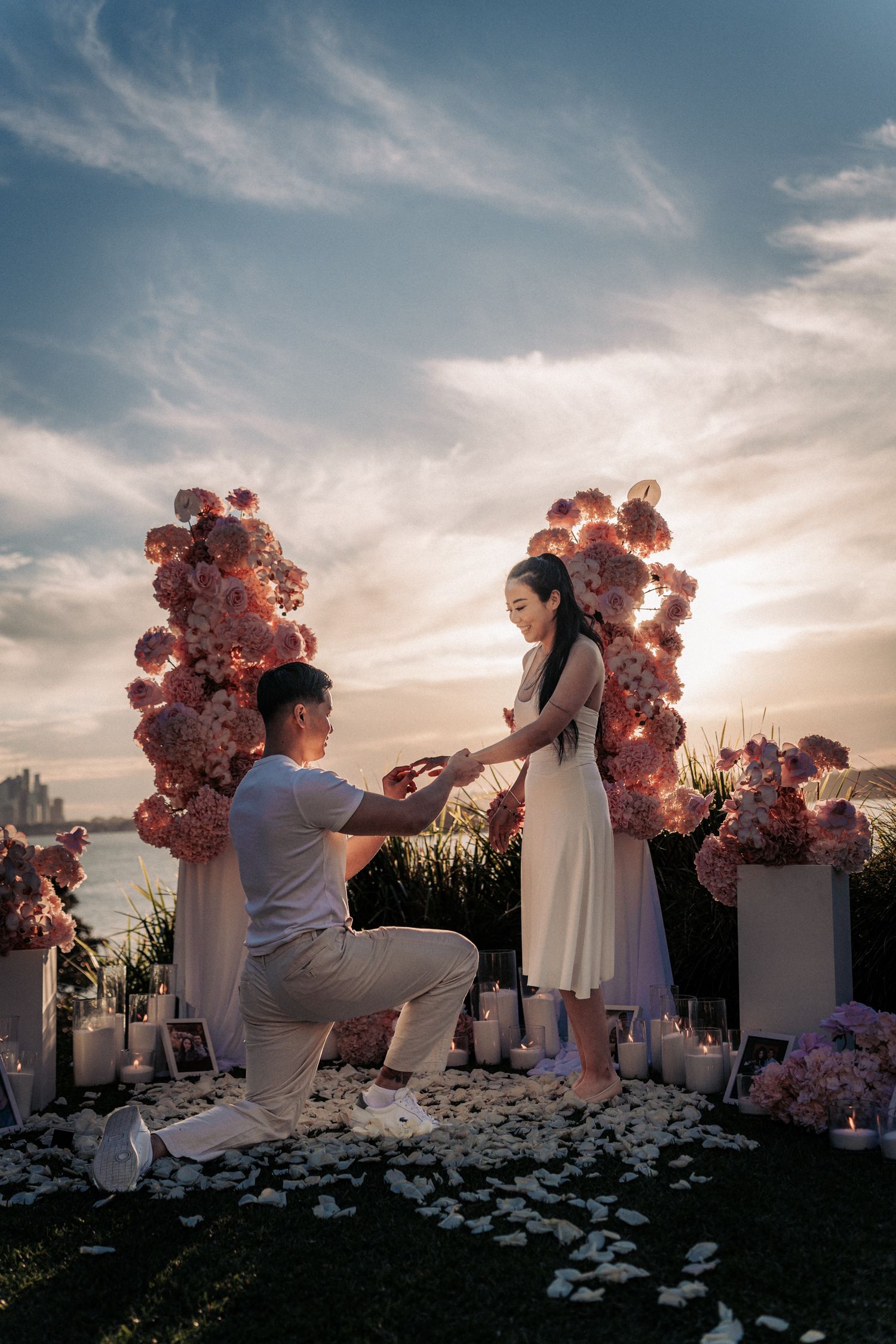 Man proposes to woman at sunset surrounded by pink floral arrangements and candles by the waterfront.
