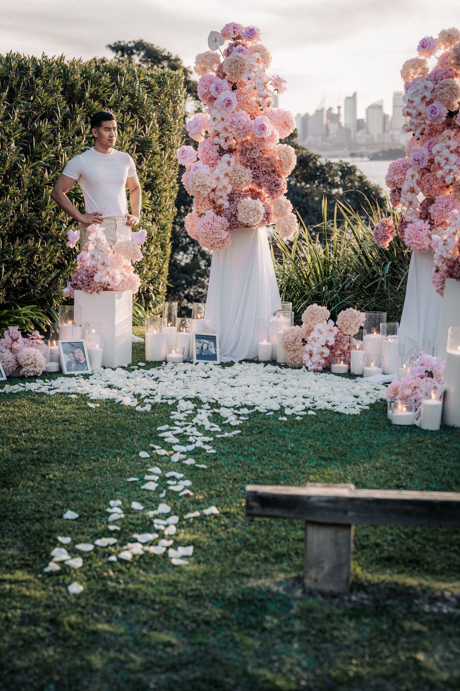 Man stands beside elegant pink floral arrangements with candles and rose petals on grass, city skyline in background.