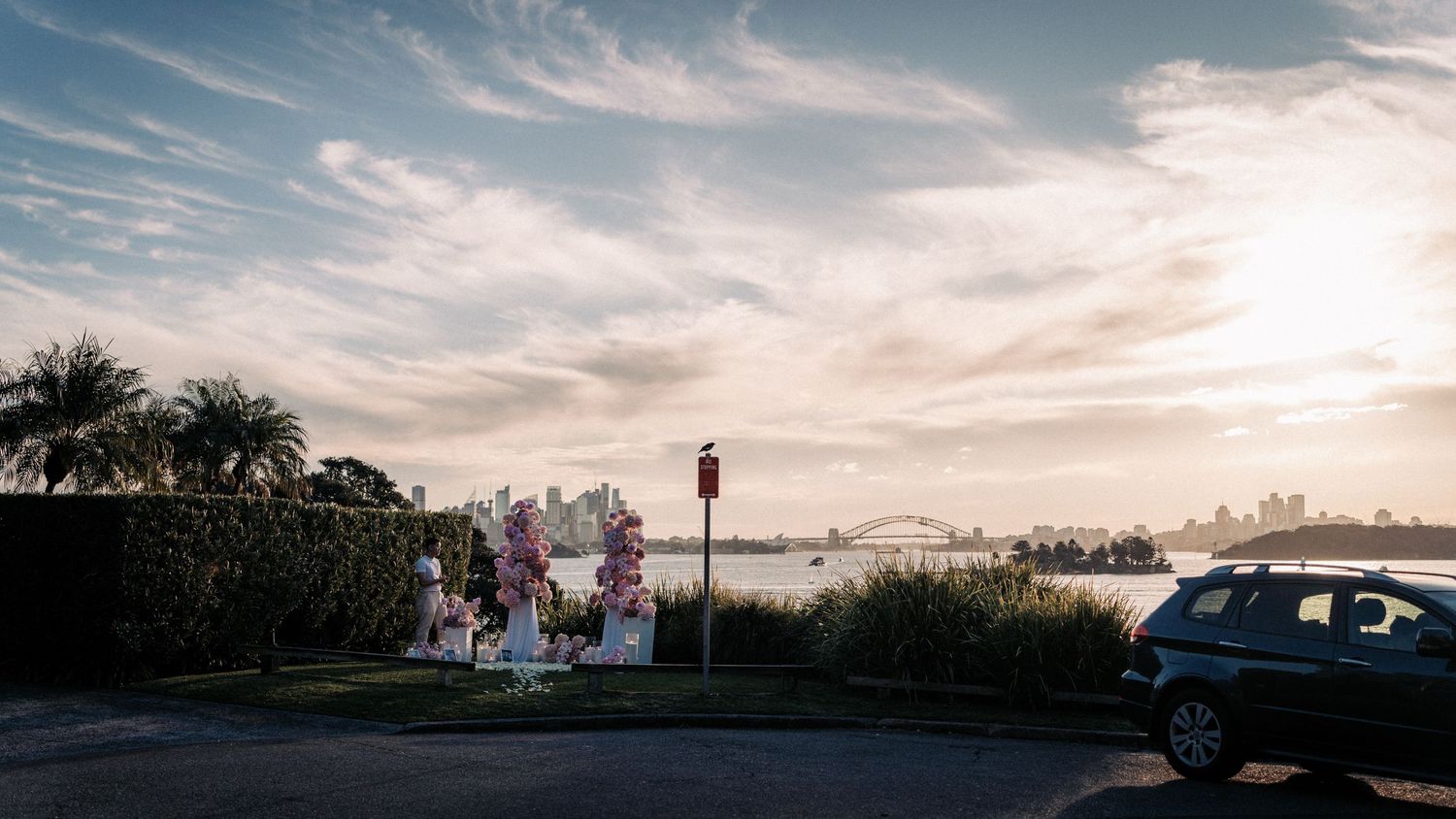 Outdoor wedding arch adorned with pink flowers overlooking Sydney Harbour Bridge at sunset with dramatic cloudy sky.