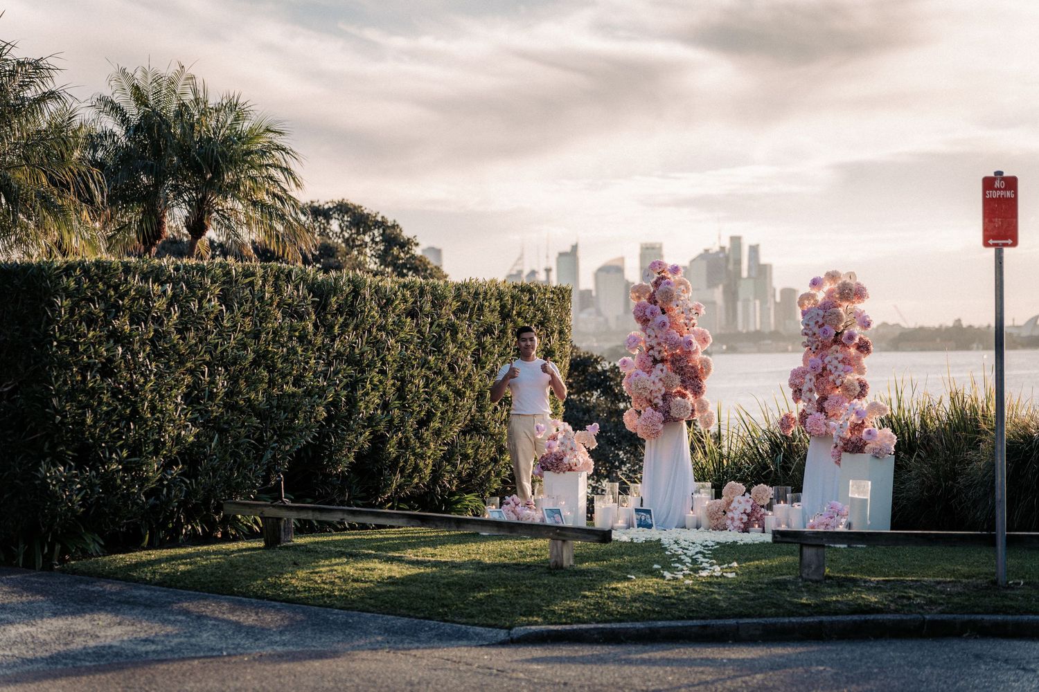 Romantic outdoor proposal setup with pink floral arrangements, candles, and city skyline backdrop near waterfront at sunset.