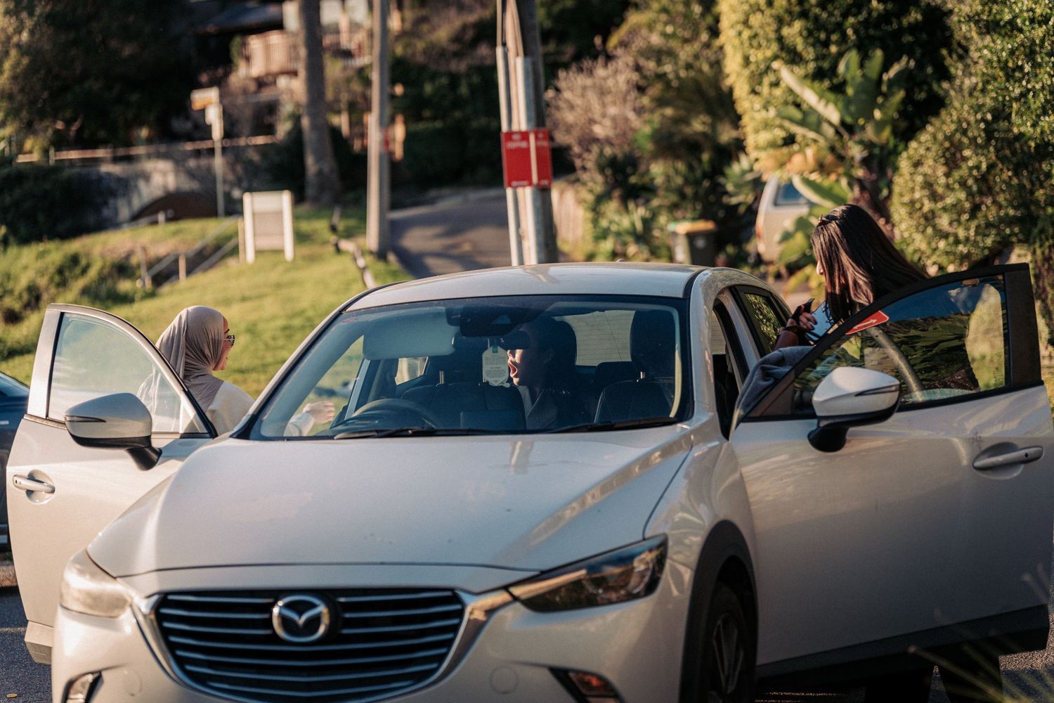 People getting into a white Mazda CX-3 car parked on a suburban street surrounded by trees.