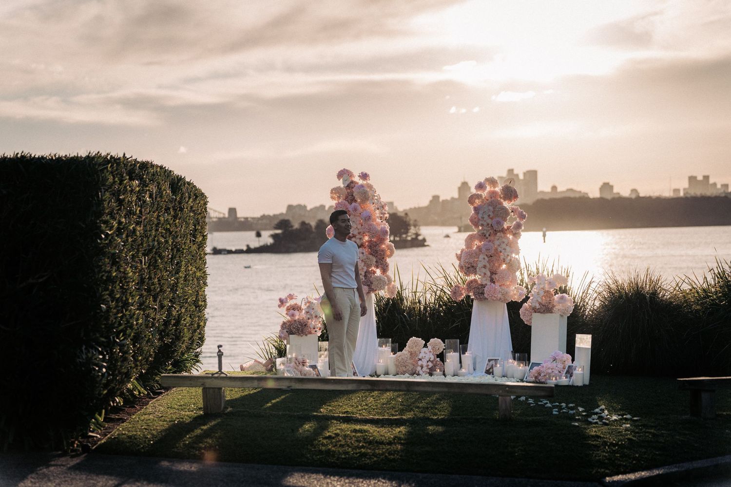 Romantic waterfront wedding setup with pink floral arrangements on white pedestals at sunset with city skyline views.
