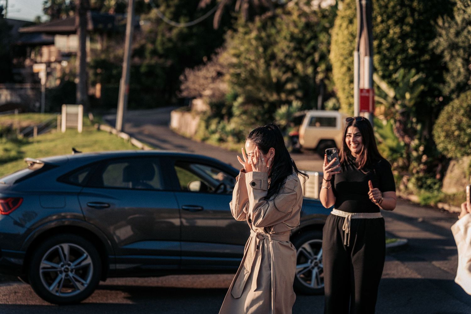 Two women standing on a suburban street near parked cars, one covering her face with hands.