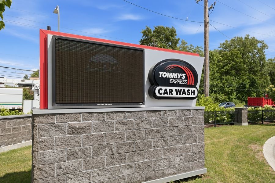 Tommy's Express Car Wash outdoor sign on stone base with digital display screen surrounded by green trees.