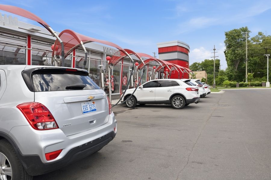 Cars lined up entering a red-roofed car wash tunnel on a sunny day, with SUVs awaiting service.