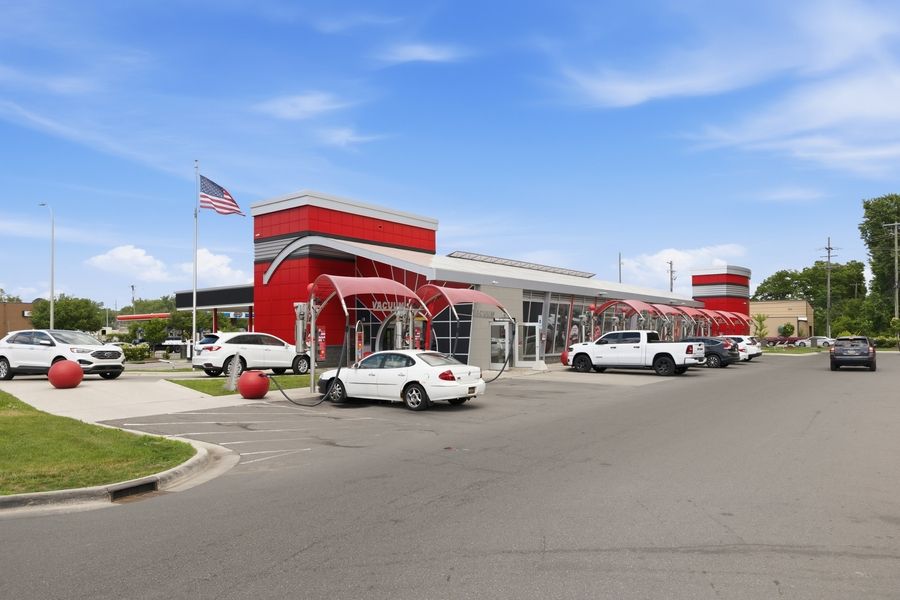 Red and white car wash facility with American flag, multiple vehicle bays, and spacious parking lot under blue sky.
