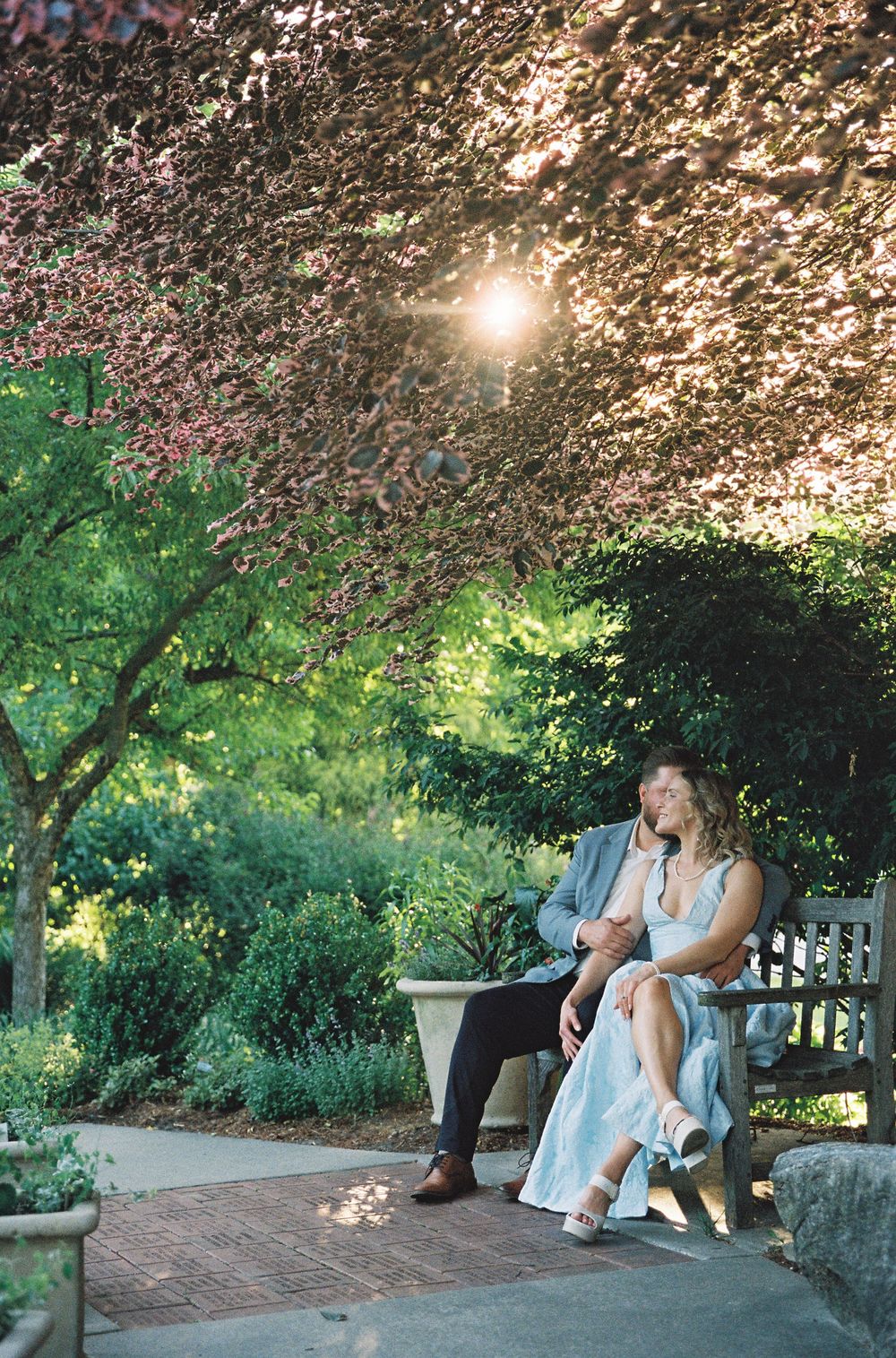 Couple sitting on a garden bench surrounded by lush greenery with sunlight filtering through flowering trees.