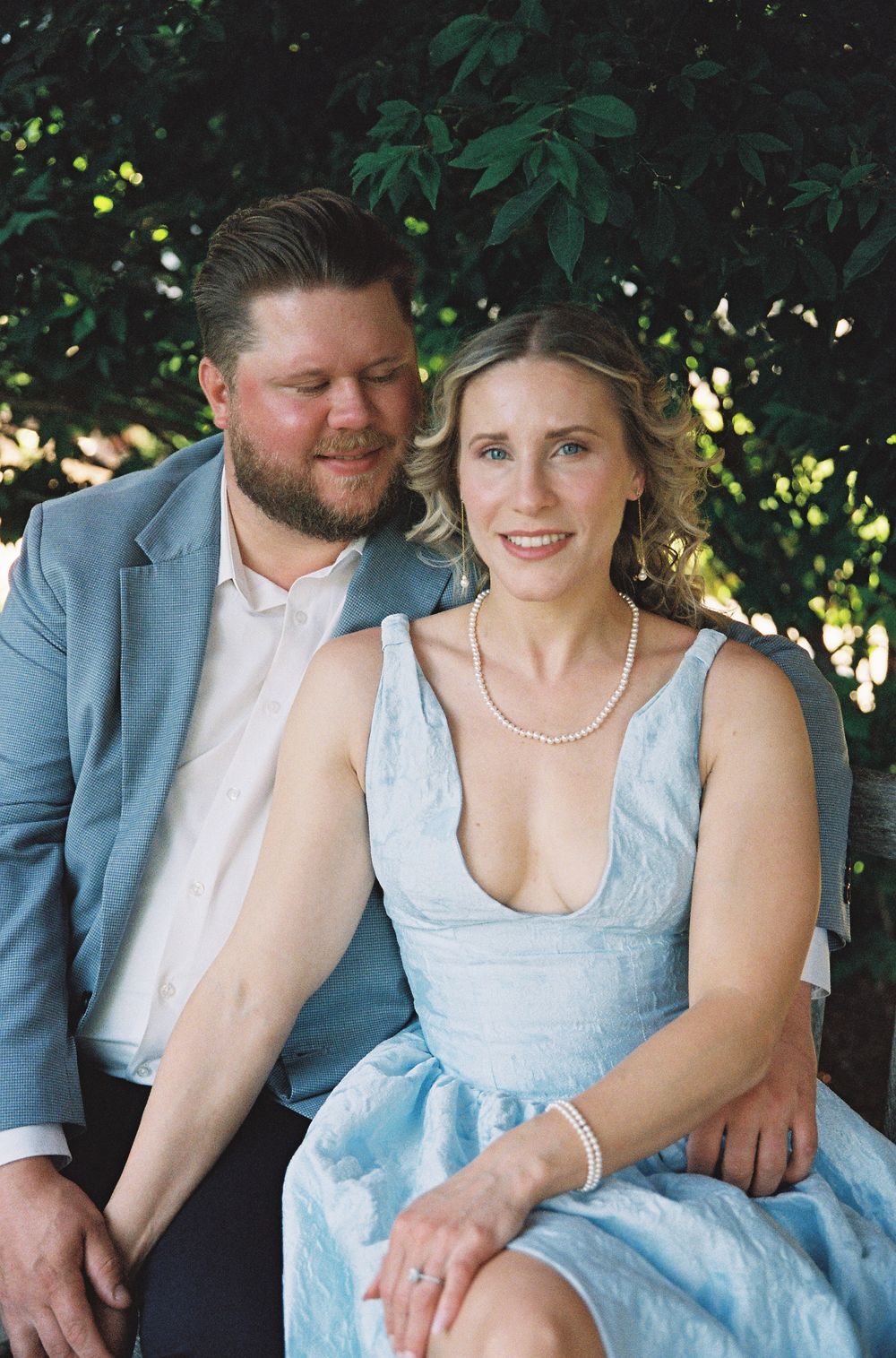 Couple in light blue formal attire sitting together outdoors, smiling, with lush green foliage background.