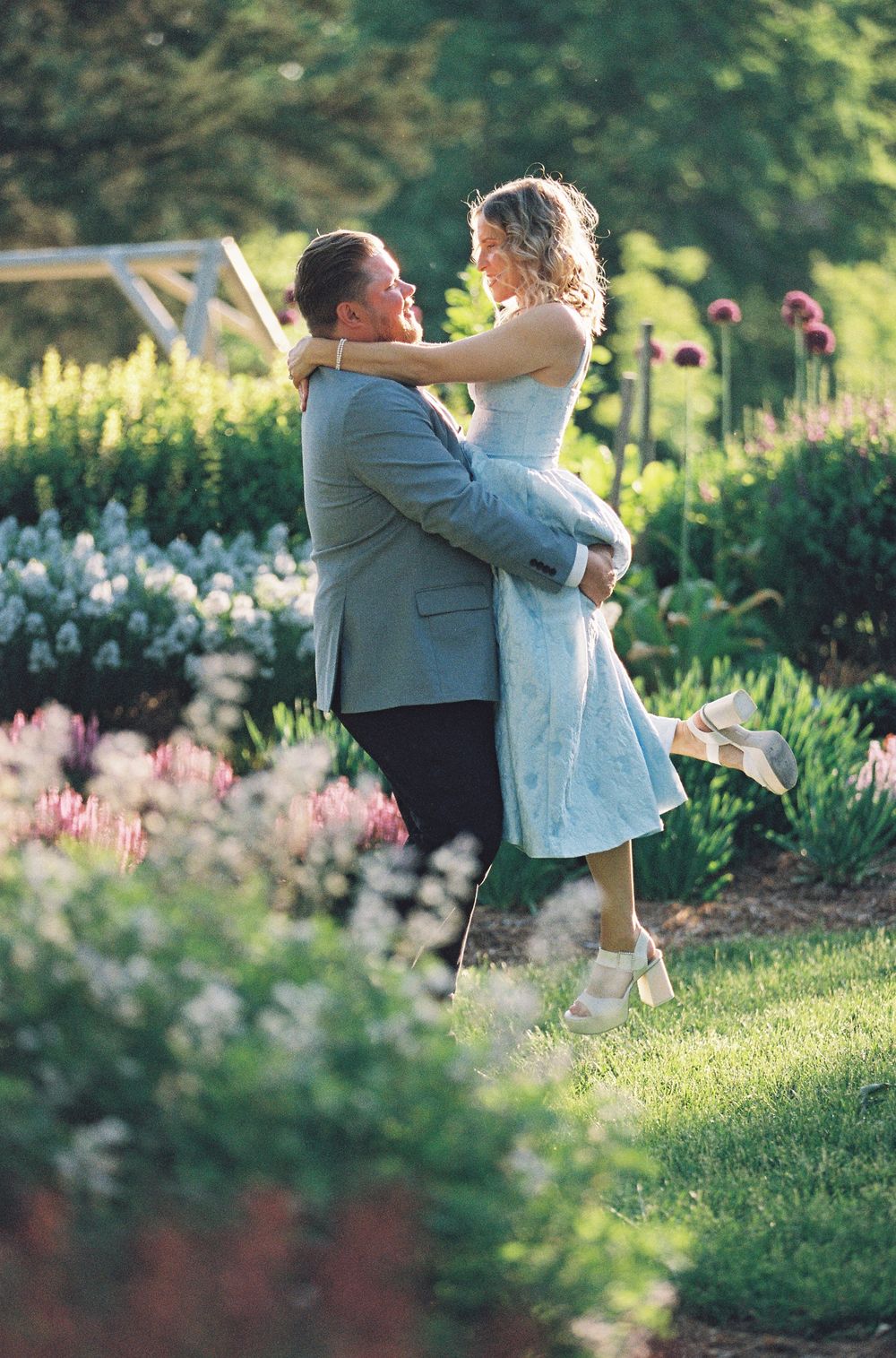 Couple embracing in a lush garden, man lifting woman in blue dress among colorful blooming flowers.