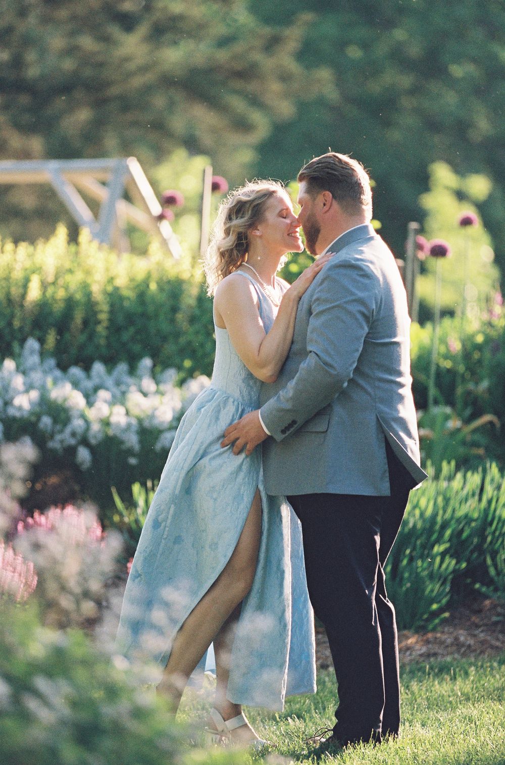 Couple sharing a romantic moment in a lush garden, woman in blue dress, man in gray suit, surrounded by blooming flowers.