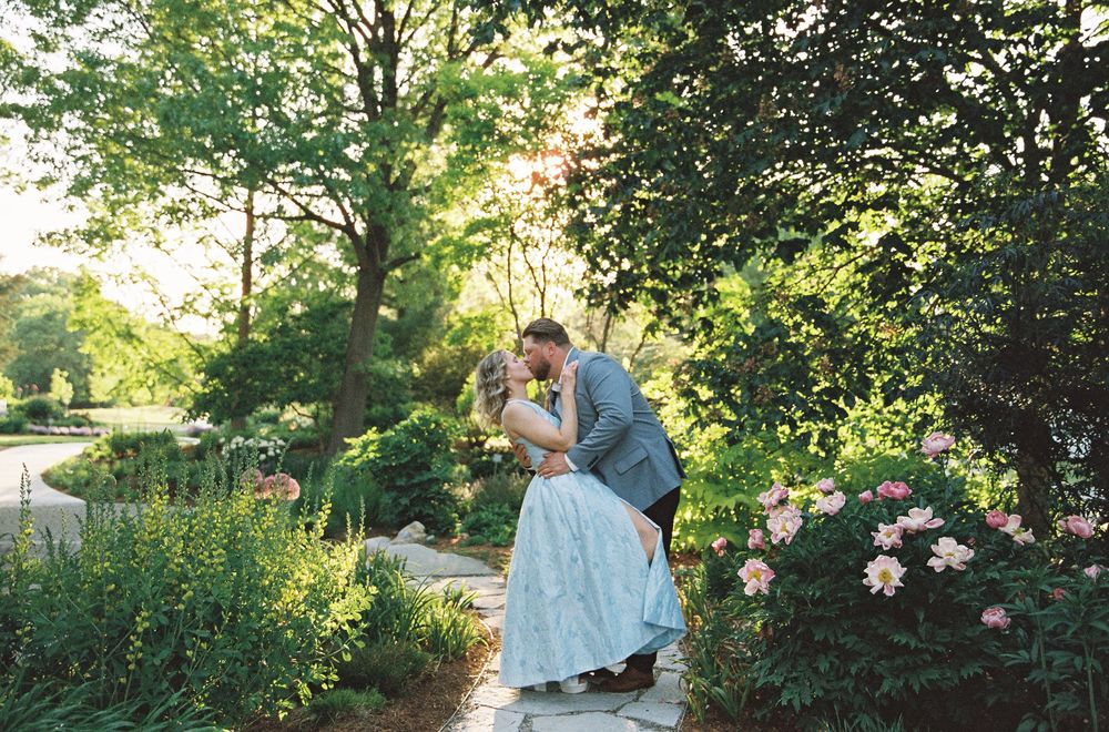 Couple sharing a romantic dip kiss surrounded by lush greenery and pink flowers in a garden wedding setting.