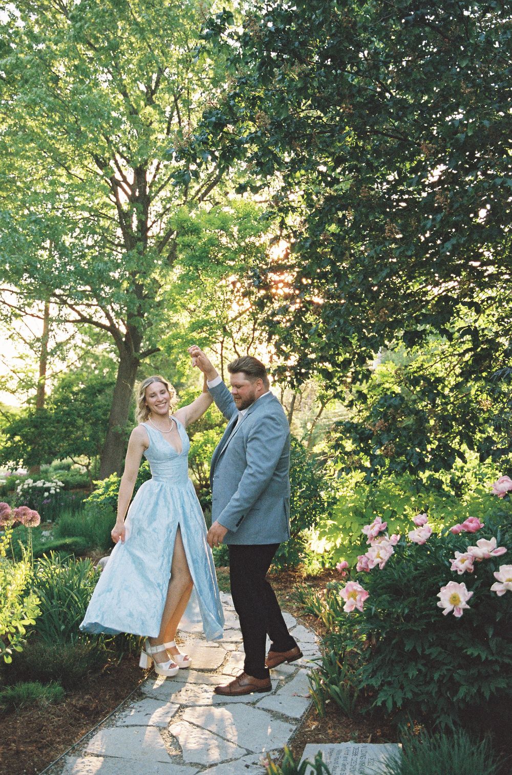 Couple dancing on a garden stone path surrounded by lush greenery and pink flowers, woman in light blue gown.