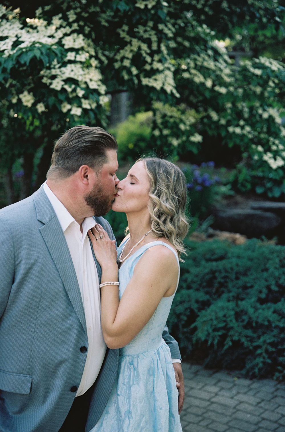 Couple sharing a romantic kiss in a lush garden, man in gray suit and woman in light blue dress.