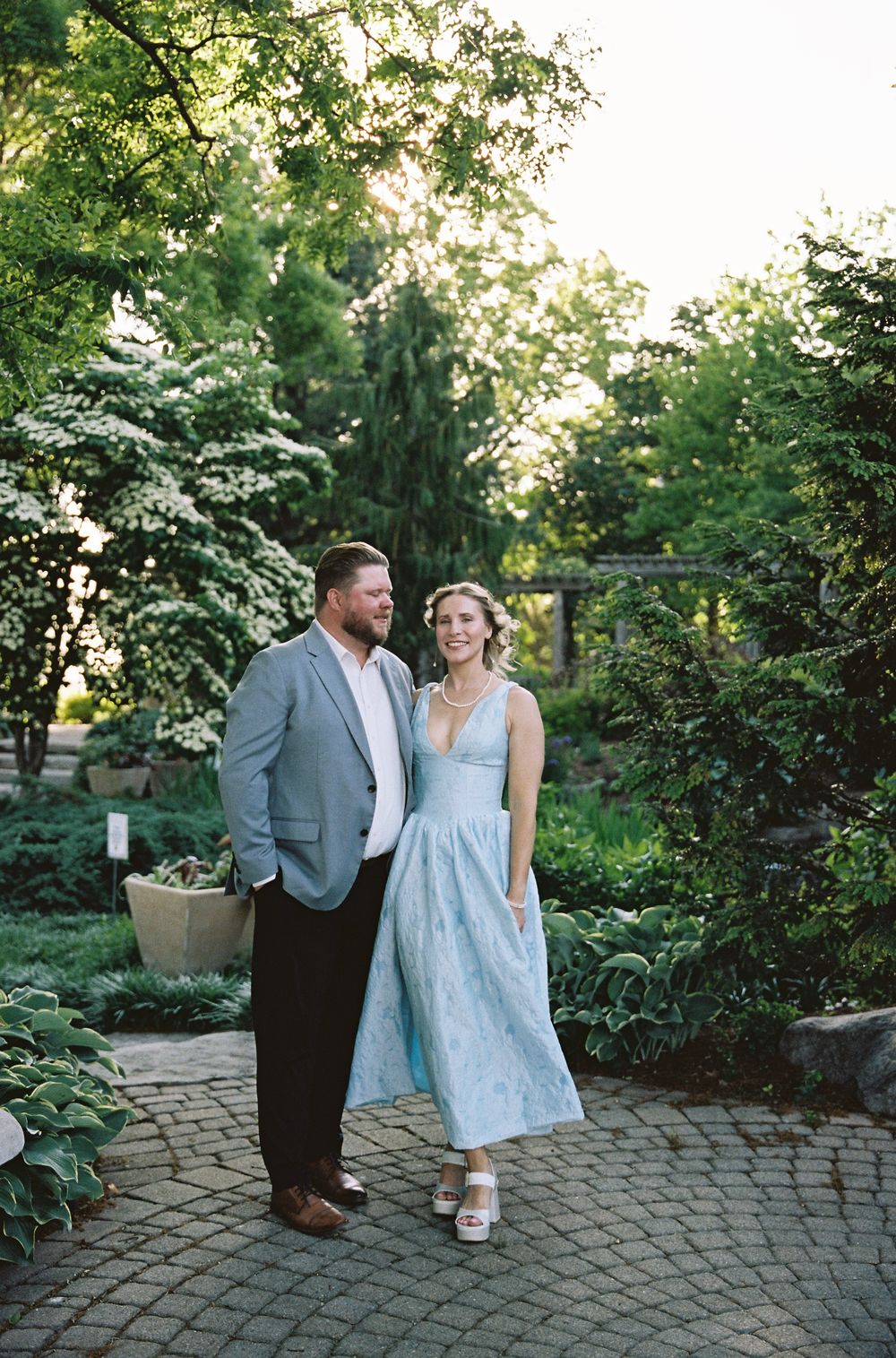 A couple smiles together on a brick garden path, woman in light blue dress and man in gray blazer, surrounded by lush greenery.