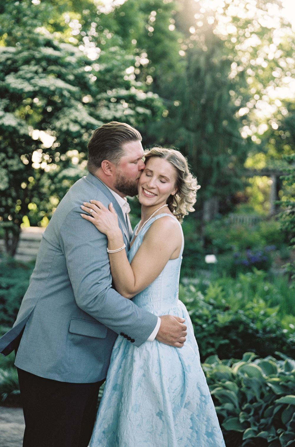Man in gray suit kissing woman in blue floral dress on cheek in romantic garden setting at sunset.