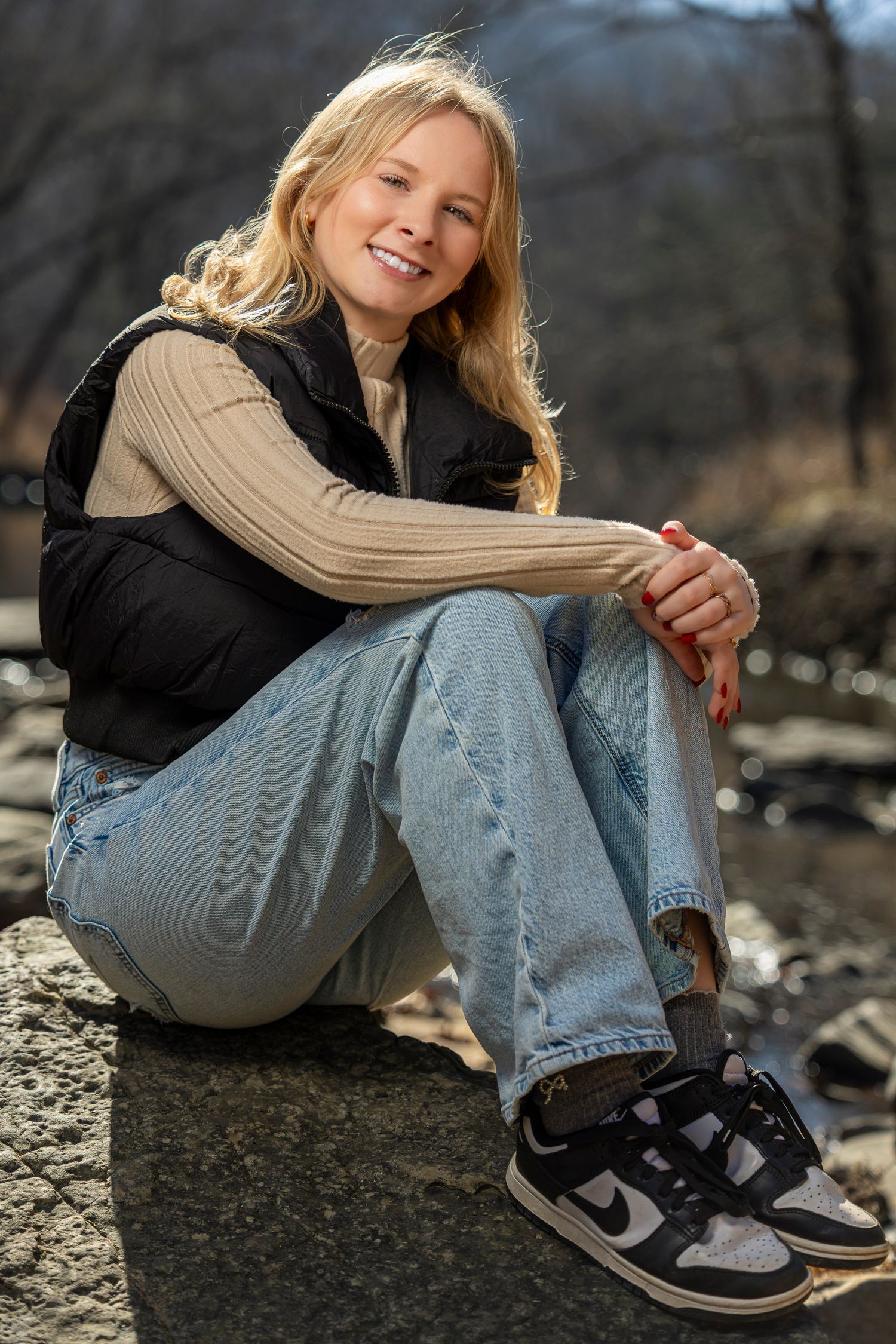Smiling blonde woman sitting on rocks outdoors, wearing a black vest, beige sweater, wide-leg jeans, and Nike sneakers.
