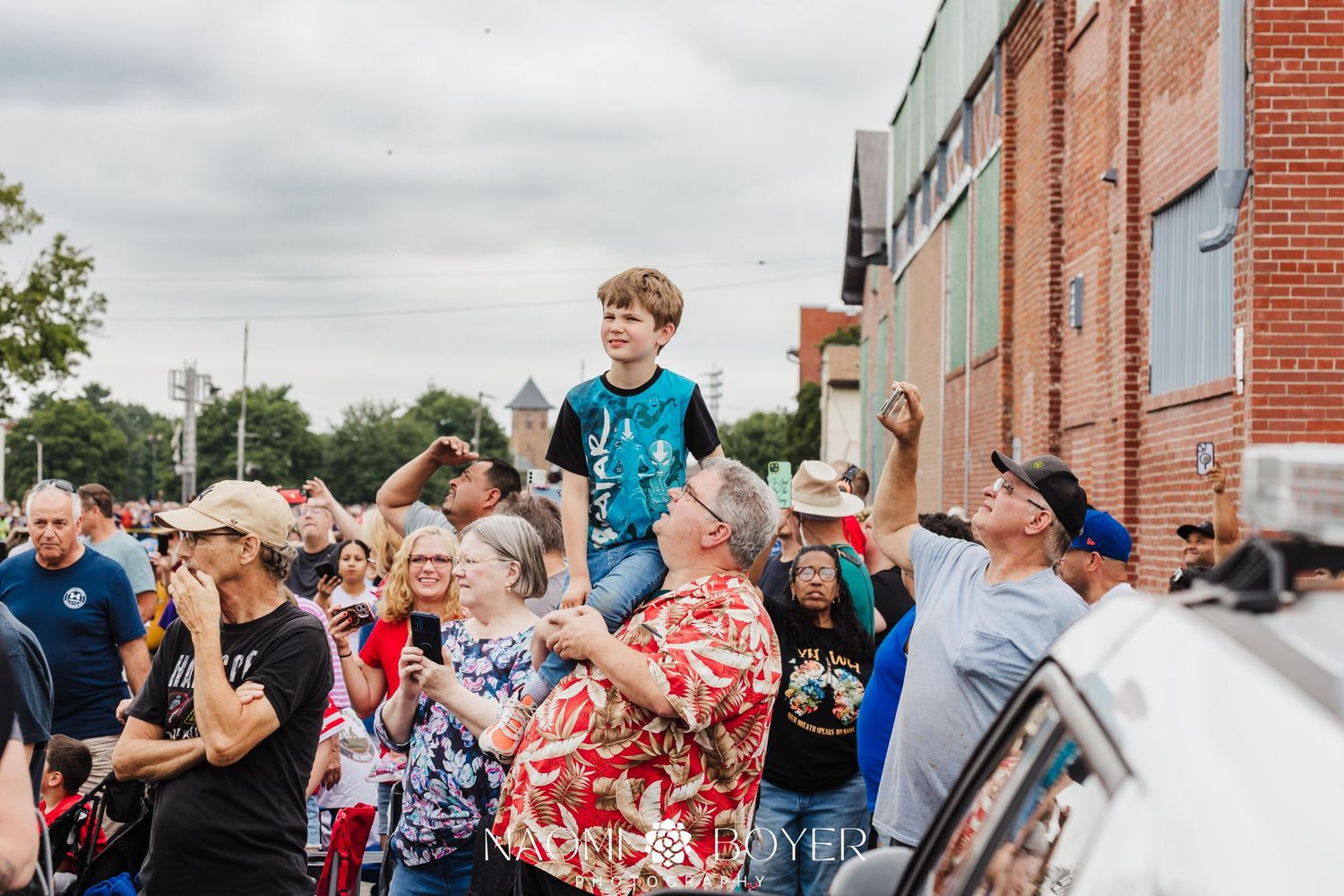 Crowd of people gathering to watch the Union Big Boy train, including a boy sitting on shoulders, cheering along a brick building street.